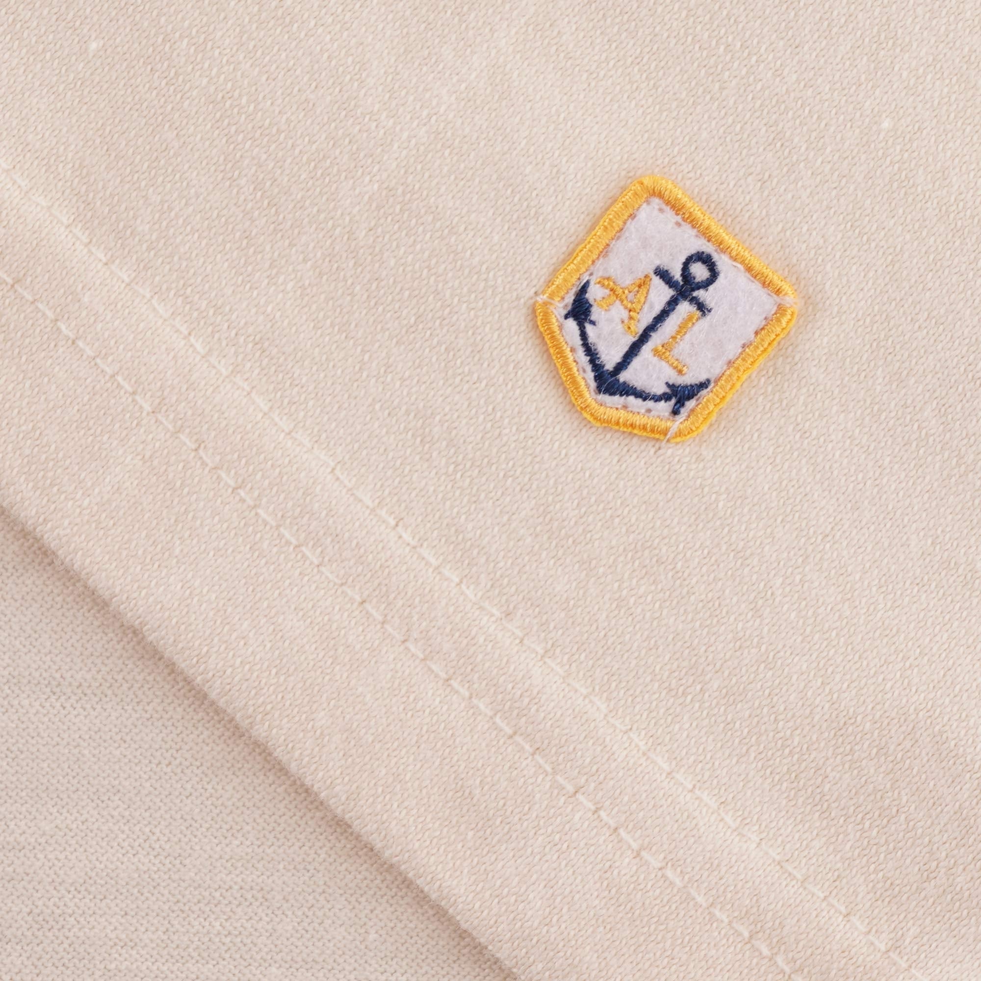 A close-up of the small woven patch featuring an anchor logo on the cream T-shirt.
