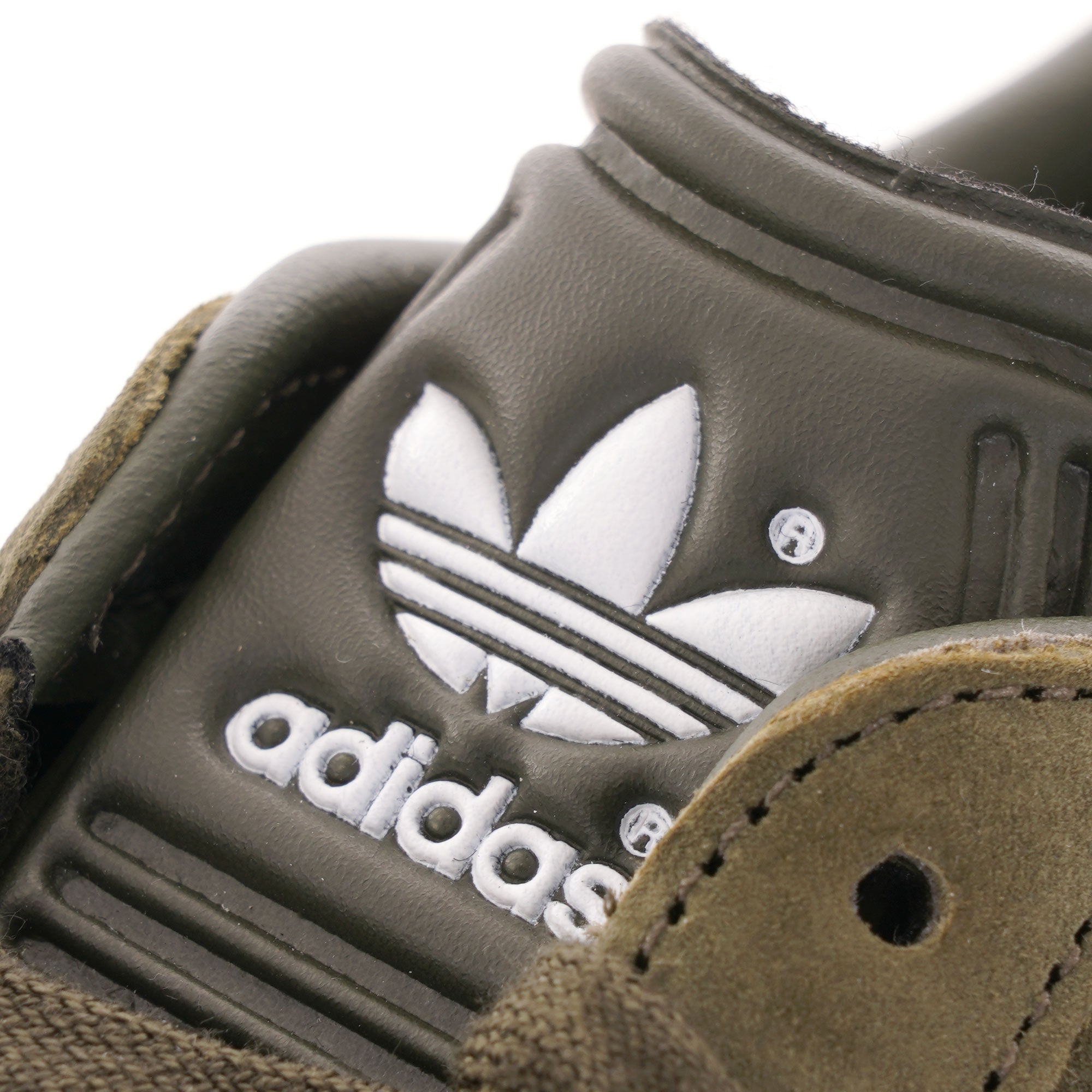 A close-up of the Adidas logo on the tongue of an olive green Gazelle trainer. 