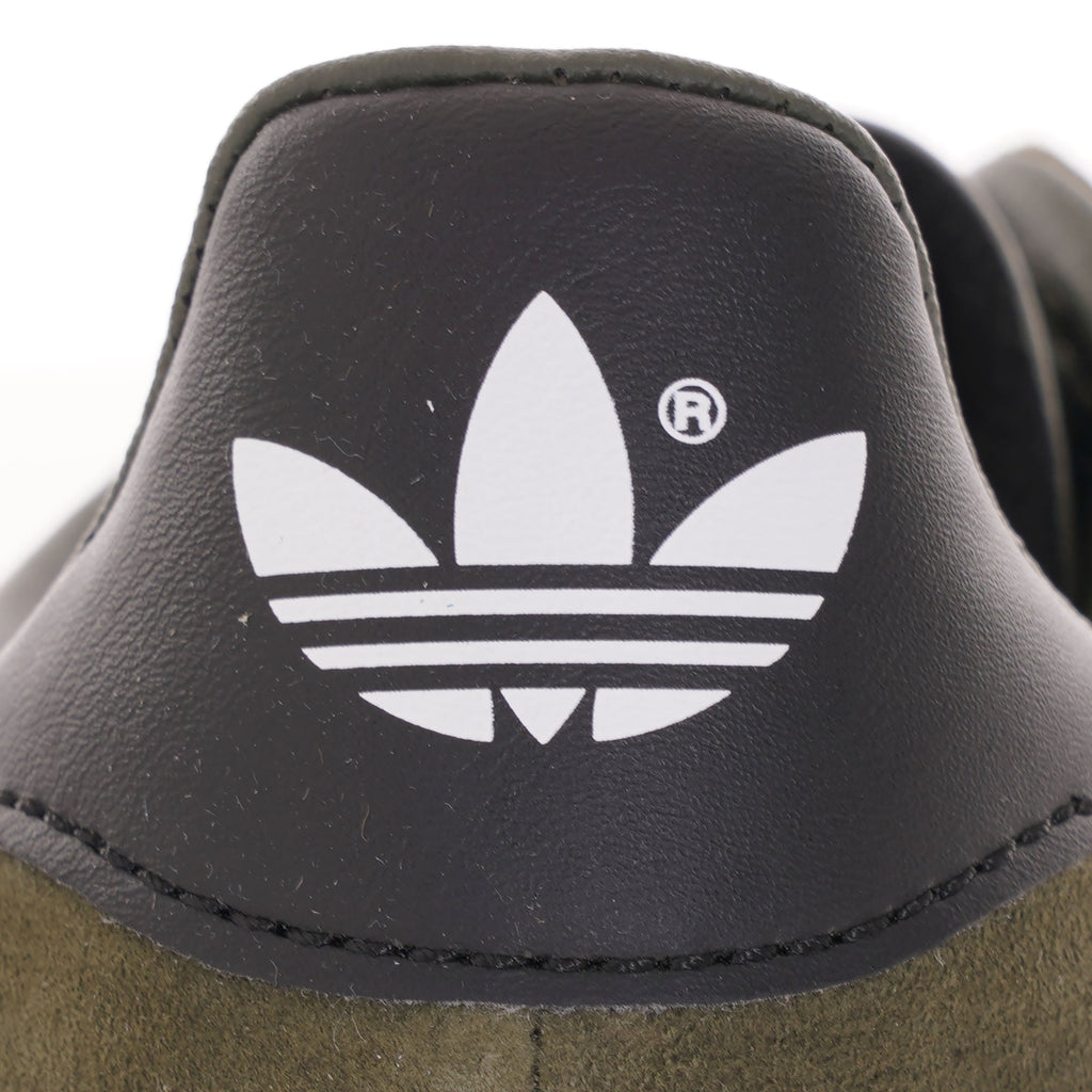 A close-up of the Adidas Trefoil logo on the black heel tab of an olive green trainer. 