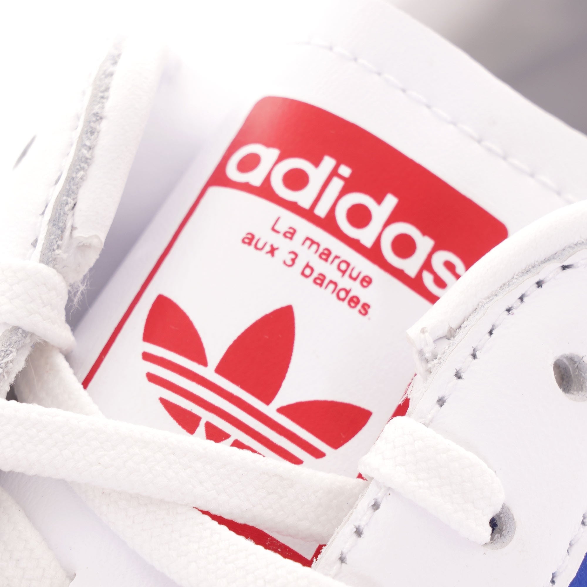 A close-up of the red Adidas logo on the tongue of a white Adidas trainer.