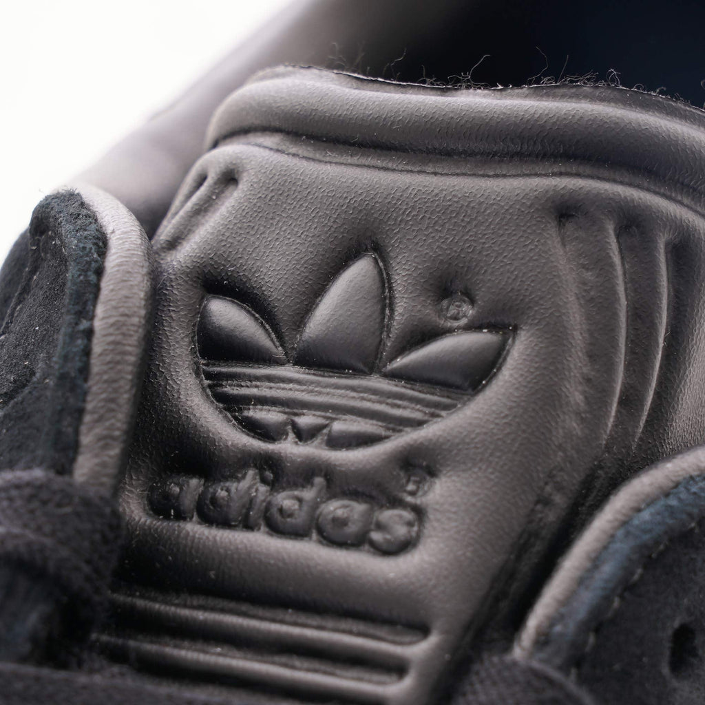 A close-up of the Adidas Trefoil logo embossed on the tongue of a black trainer.