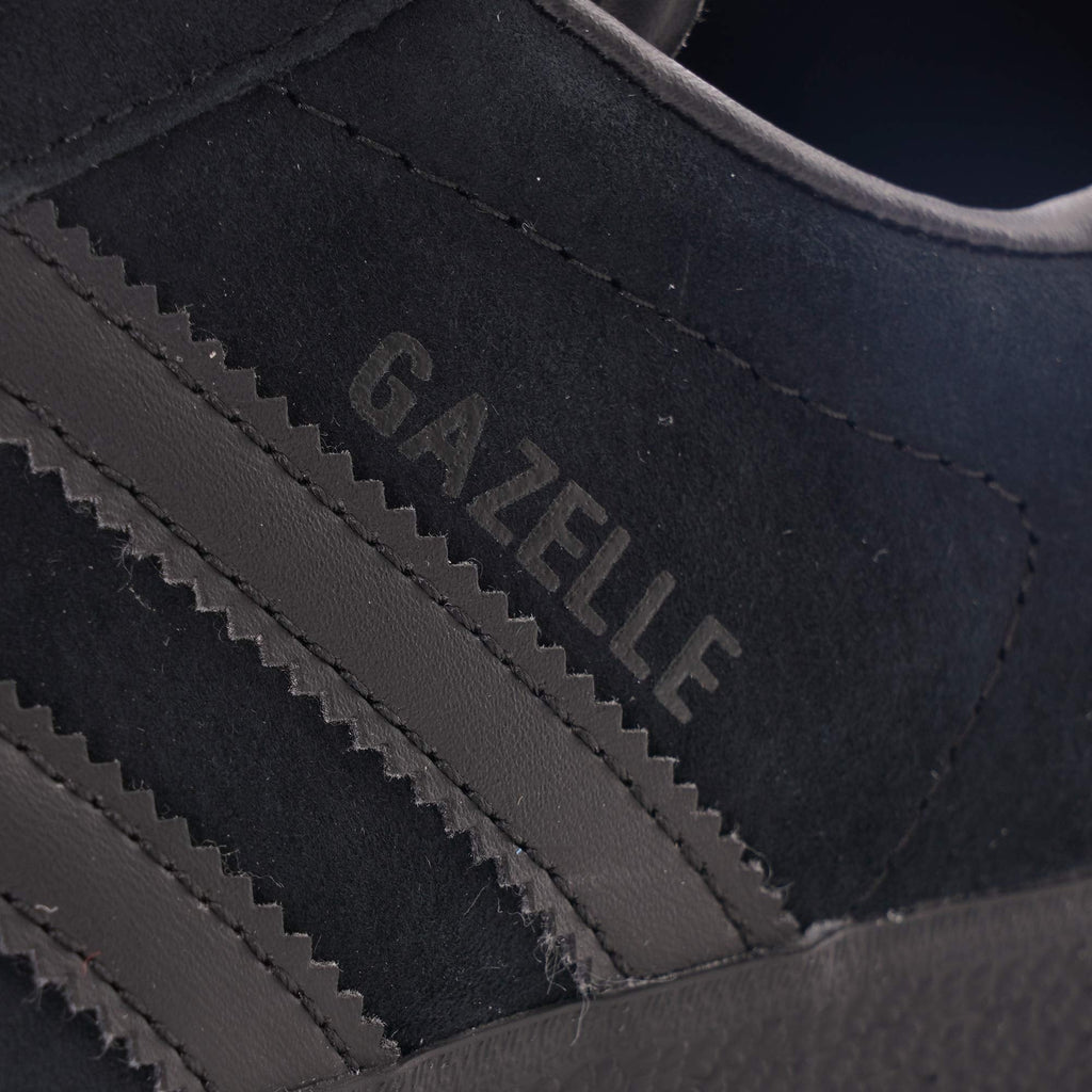 A close-up of the 'Gazelle' branding on the side of a black Adidas trainer.