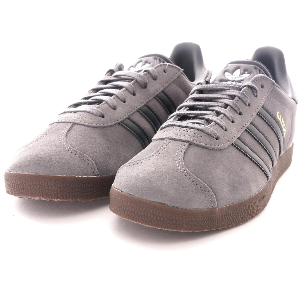 A pair of grey Adidas Gazelle trainers with black stripes and brown soles, viewed from an angle.