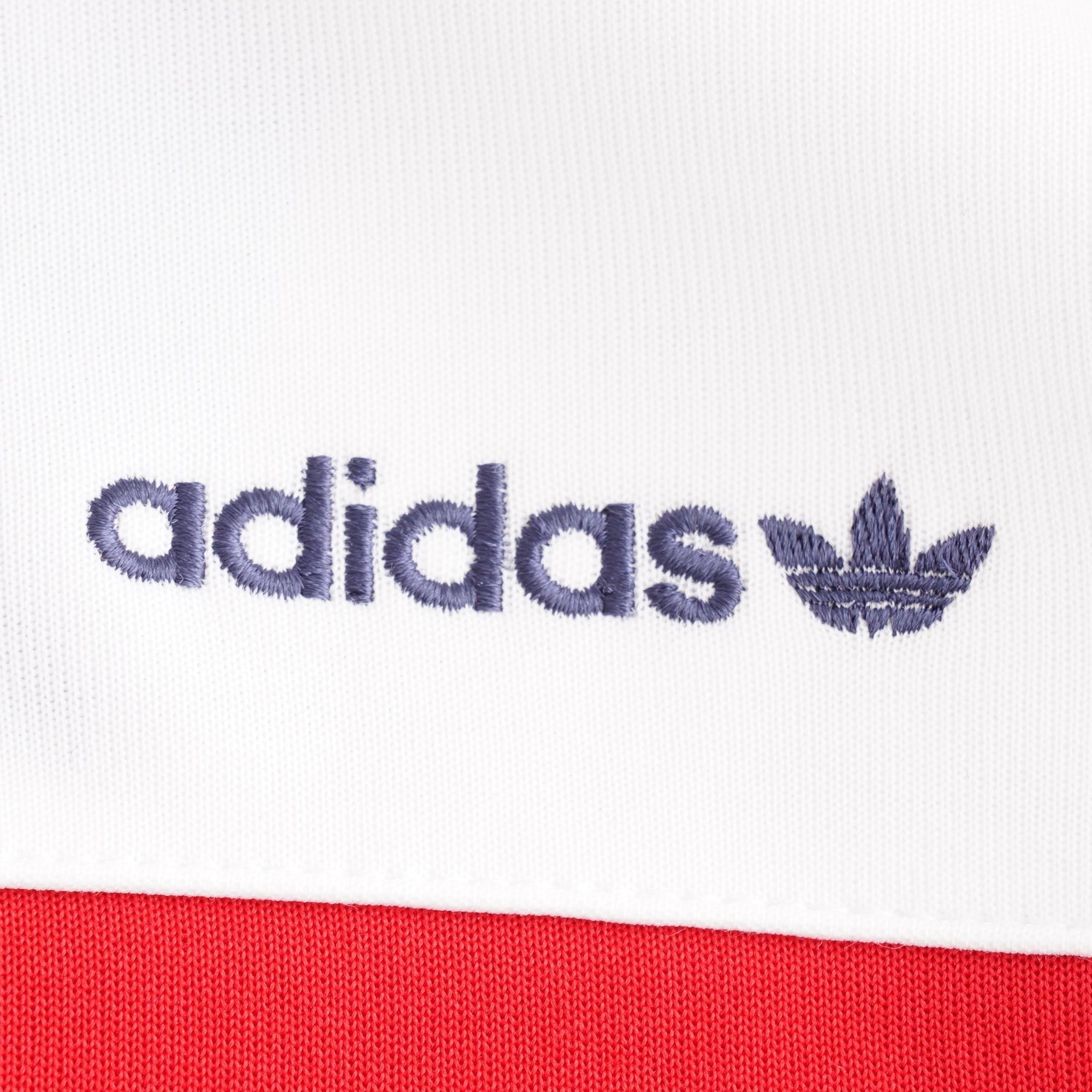 A close-up of the embroidered navy Adidas logo on the white fabric of the track top.